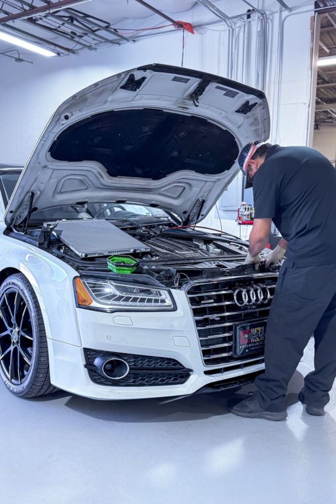 Technician in black overalls and a US flag headband works under the open hood of a white Audi in a bright workshop