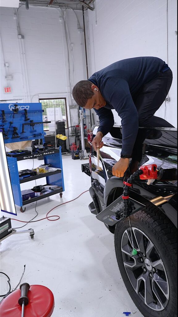 Mechanic crouches at the rear wheel of a black SUV in a bright auto shop, using a tool mounted near the fender.