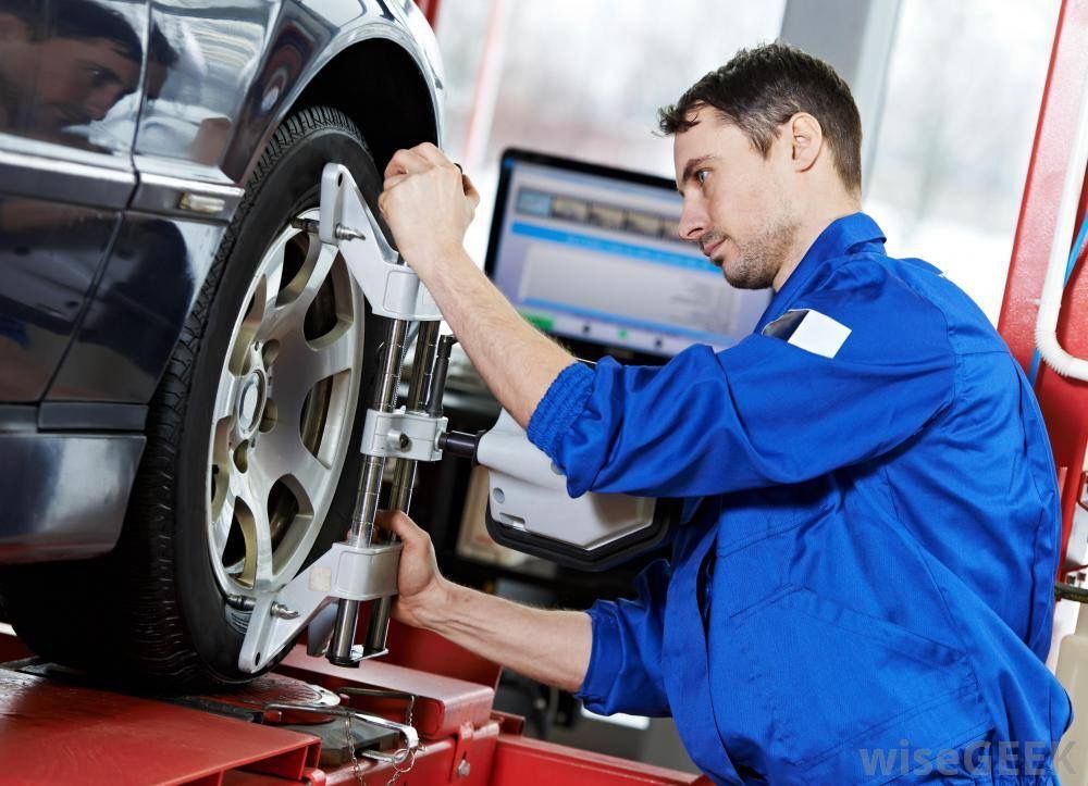 Auto technician in blue overalls works on a car wheel using a wheel-alignment apparatus on a raised vehicle, with a diagnostic screen nearby.