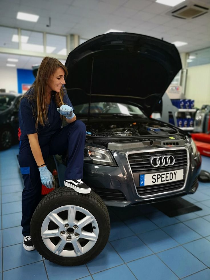 Female mechanic in blue scrubs and gloves lifts a tire beside a dark Audi with the hood up in a repair shop.
