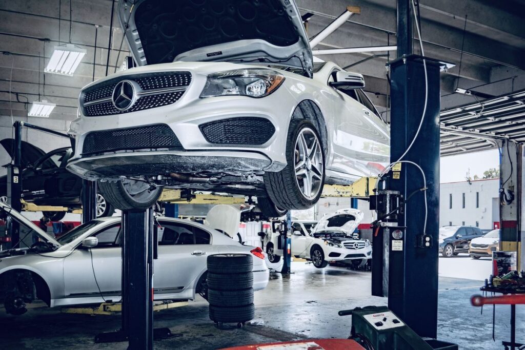 Silver Mercedes SUV on a lift with its hood raised in a busy auto repair shop setting.