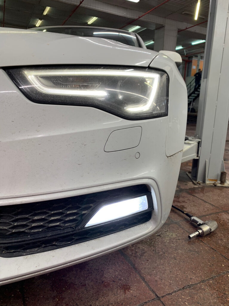Close-up of a white car's front end in a workshop, with LED headlight and daytime running light illuminated, and a tool lying on the floor nearby.