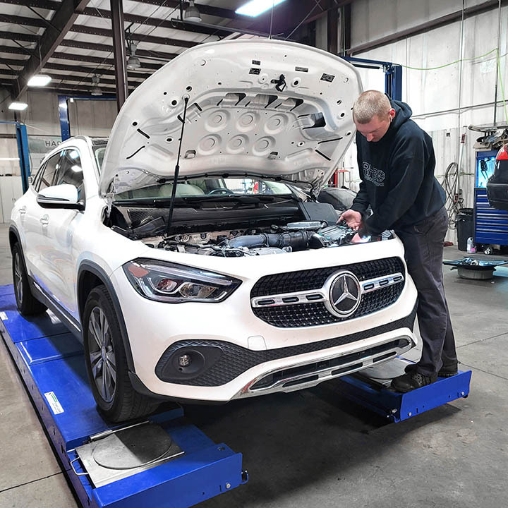White Mercedes-Benz SUV on a blue service lift with the hood open, a mechanic inspecting the engine in a workshop.