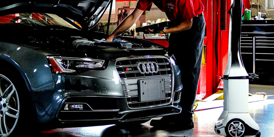Mechanic inspecting the engine under the hood of a dark gray Audi in a car repair shop.