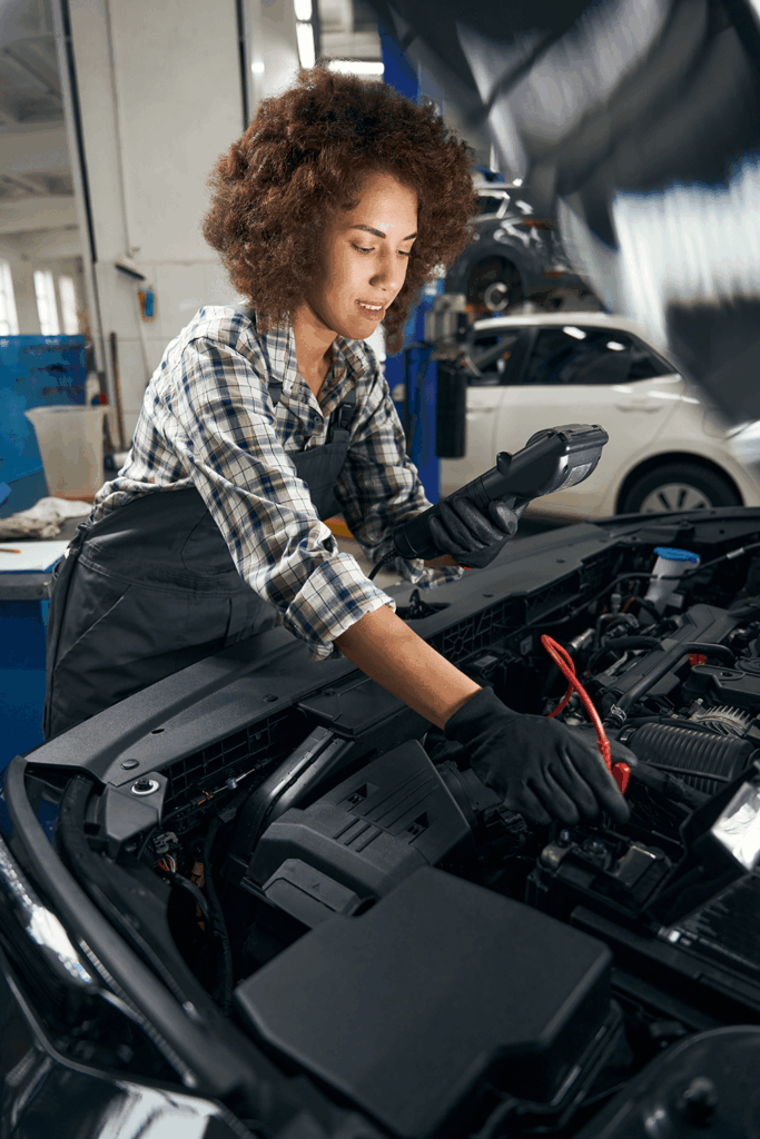 Female mechanic in a plaid shirt and overalls using a handheld diagnostic tool on a car engine in a repair shop