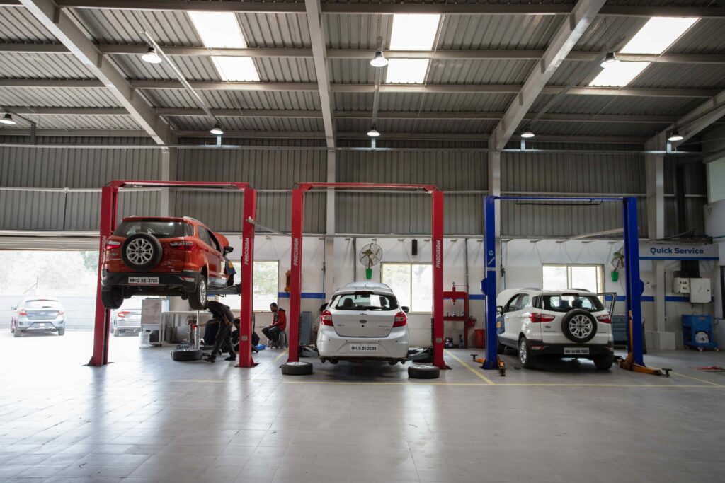 Red SUV is lifted on a hydraulic car lift inside a spacious auto repair shop with white sedans on the ground and technicians at work in the background of a high-ceilinged workshop.