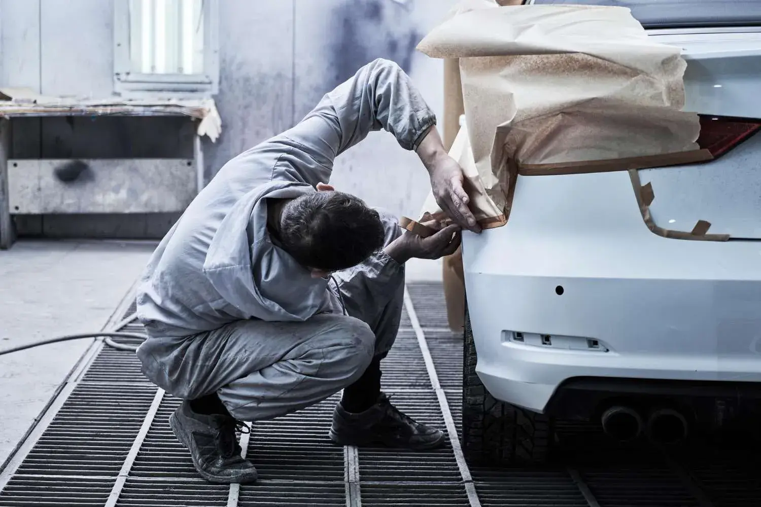 Auto body worker kneeling by a white car, masking the rear bumper with brown paper for painting in a shop.