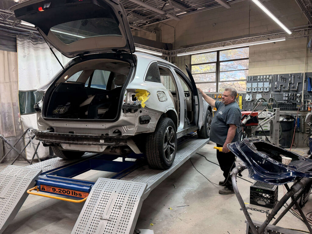 Silver SUV on a car lift in a repair shop with rear hatch open and technician inspecting the side door area