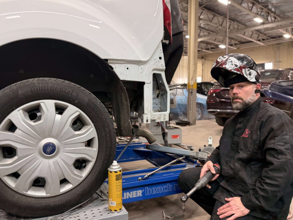 A mechanic in a skull-pattern helmet and black coveralls kneels beside a white van lifted on a hydraulic lift in a busy auto shop, tools nearby.