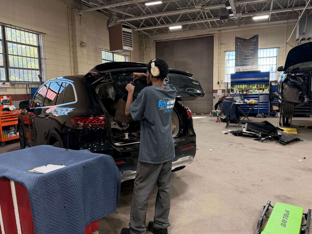 Tech wearing headphones works at open tailgate of black SUV in an auto shop, using tools on the rear interior panel as other car parts lie nearby.