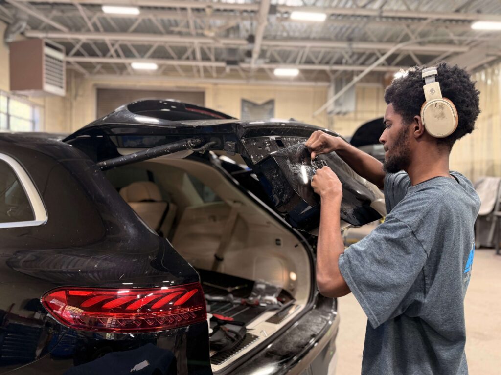 Technician wearing beige over-ear headphones works on the rear window area of a black SUV inside an auto repair shop.