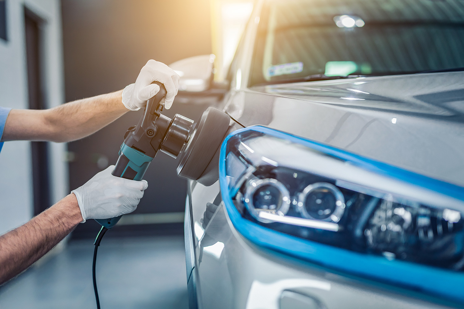 Technician wearing gloves buffing a car's front fender with a power polisher in a workshop.