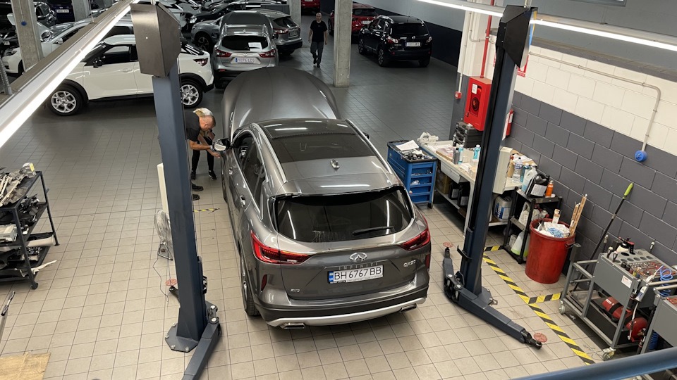 Silver SUV on a two-post lift in a well-lit auto repair shop, mechanic inspecting the left front area with the hood open.