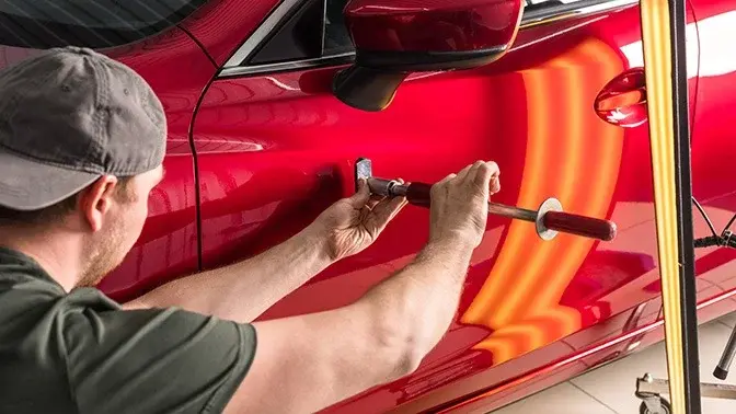 Auto body technician repairing a red car door with a hammer and punch in a workshop.