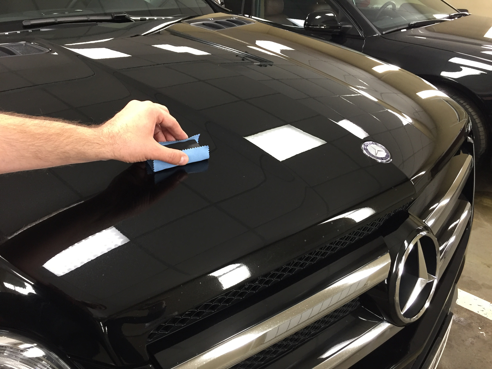 Person cleaning a glossy black car hood with a blue microfiber cloth, Mercedes emblem visible nearby in a garage setting