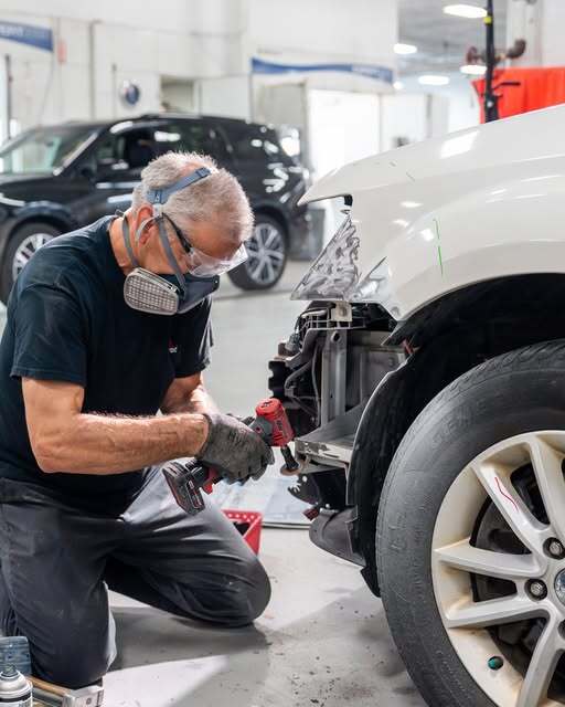 Mechanic wearing a respirator mask and safety glasses kneeling and repairing the front of a car in a workshop.