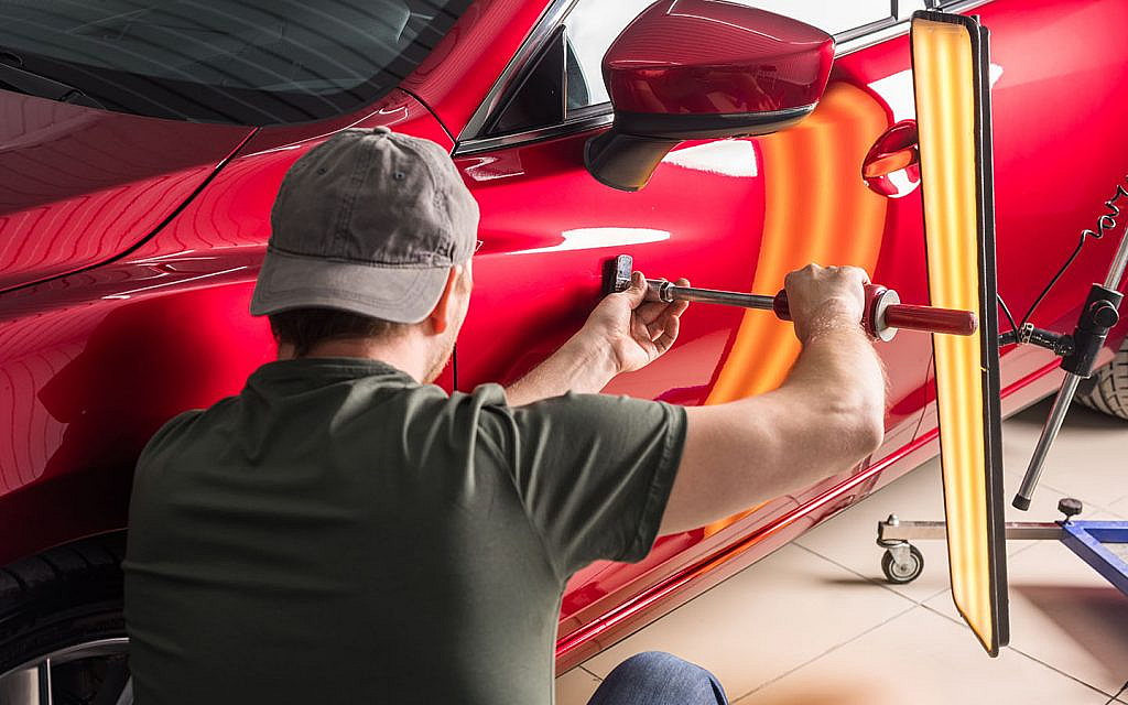 Mechanic wearing a cap tightens a tool against the red car's door in a workshop, with equipment nearby.