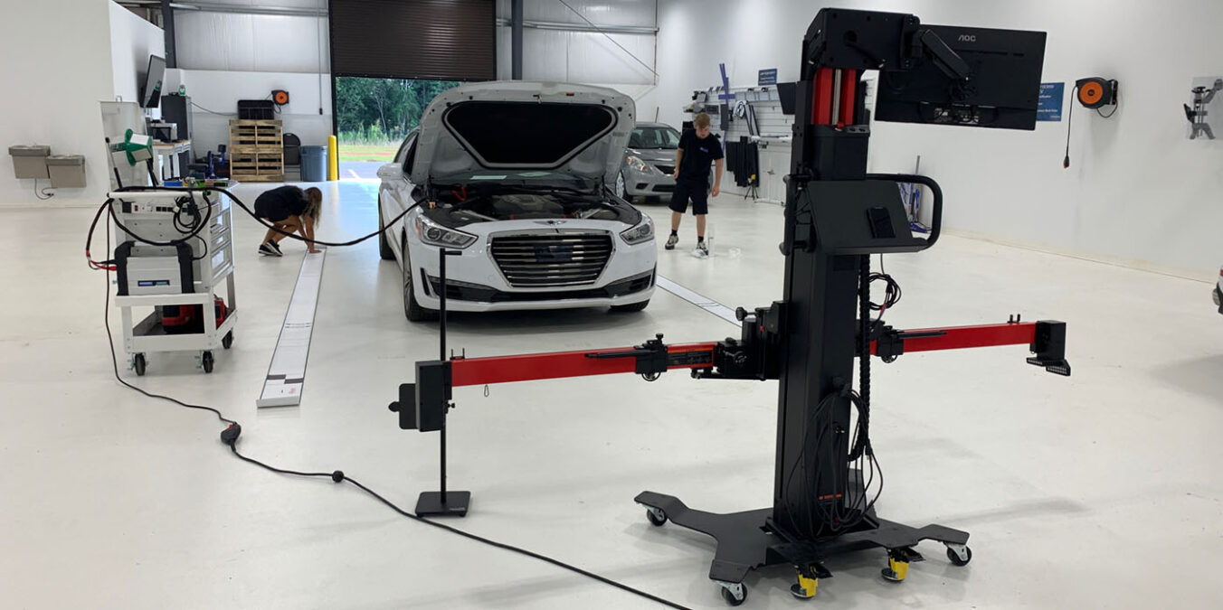 White sedan with hood open on a diagnostic/rig system in a bright workshop; two technicians nearby unfold the scene of testing or calibration.