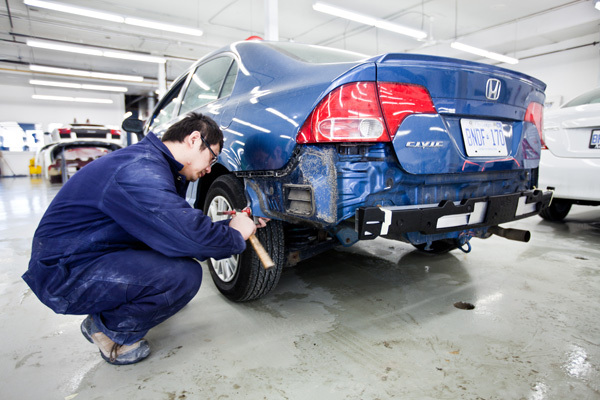 Auto mechanic crouched beside the rear wheel of a blue Honda Civic in a workshop, using a tool to work on the wheel.