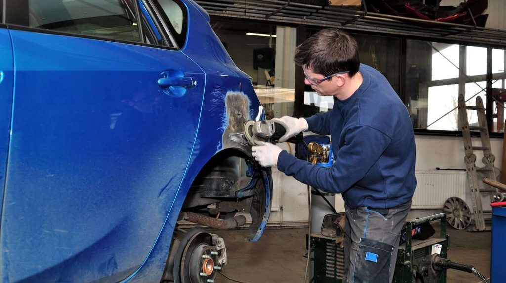 Male mechanic wearing gloves and safety glasses grinding a dent on a blue car in a repair shop.