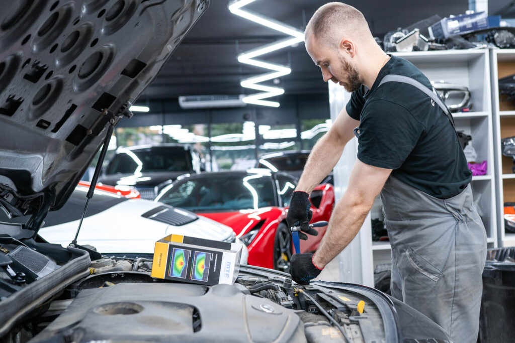 Male mechanic in gray coveralls leaning over a car engine with hood open in a modern auto shop.