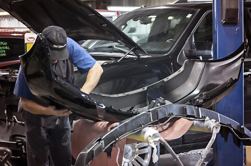 Auto mechanic in a blue shirt and cap removes a black bumper from a car on a lift in a busy repair shop with the hood open nearby.
