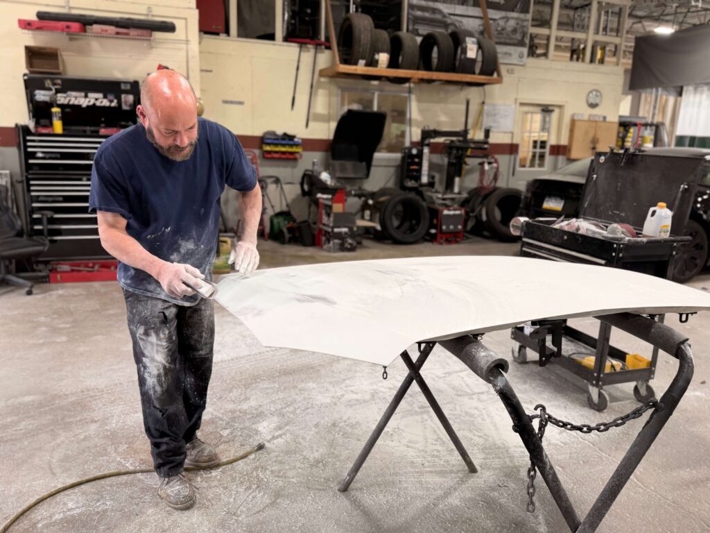 Man in a blue shirt sands a large white curved surface on a work table in a garage workshop with tools and tires in the background.