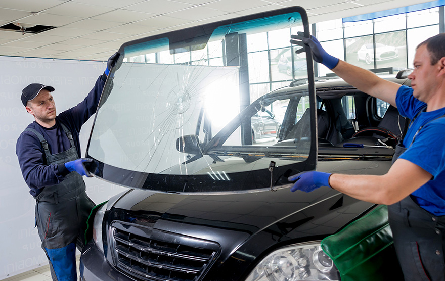Two auto technicians wearing gloves install a cracked windshield into a black SUV in a workshop.