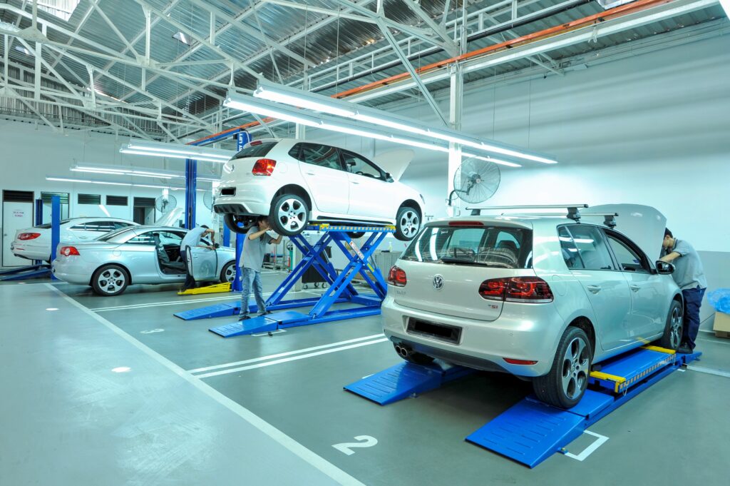 Auto repair shop with a white car on a raised lift and a mechanic inspecting under the hood in bright workshop lighting.