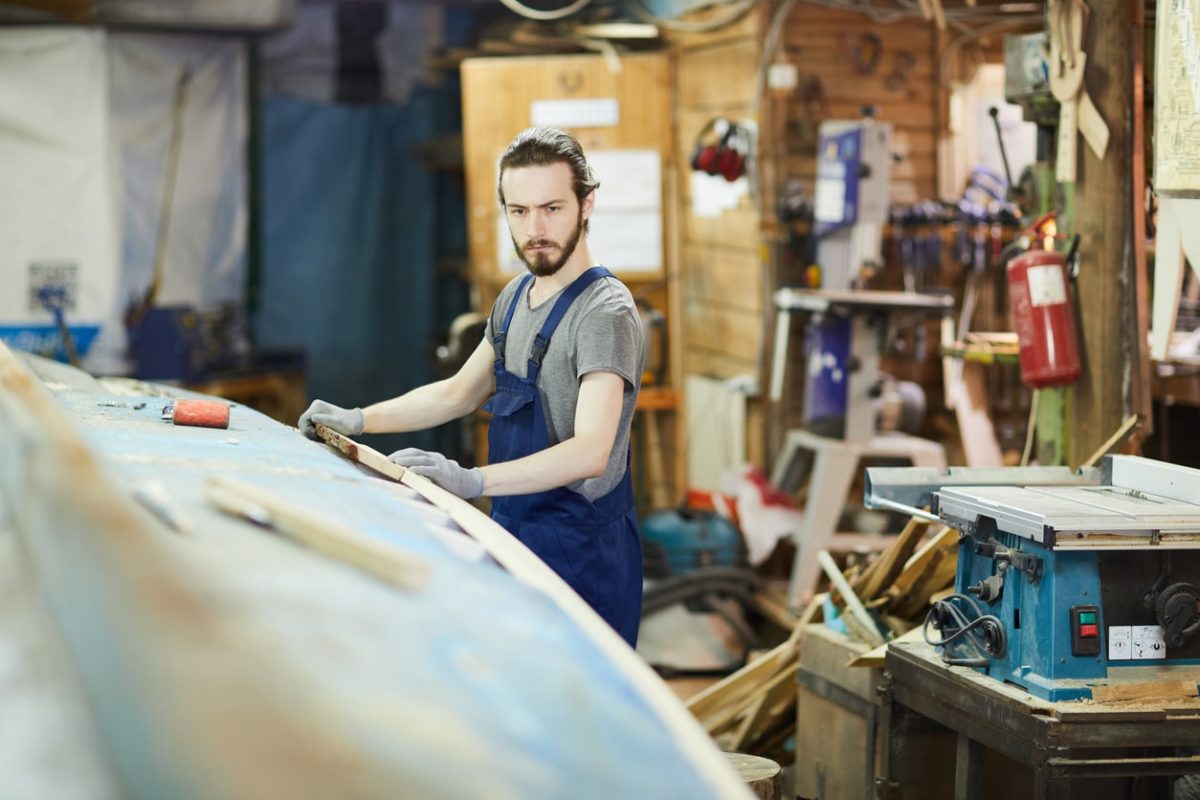 Carpenter in blue overalls planes a curved wooden surface in a cluttered workshop, wearing gloves and a gray t-shirt.
