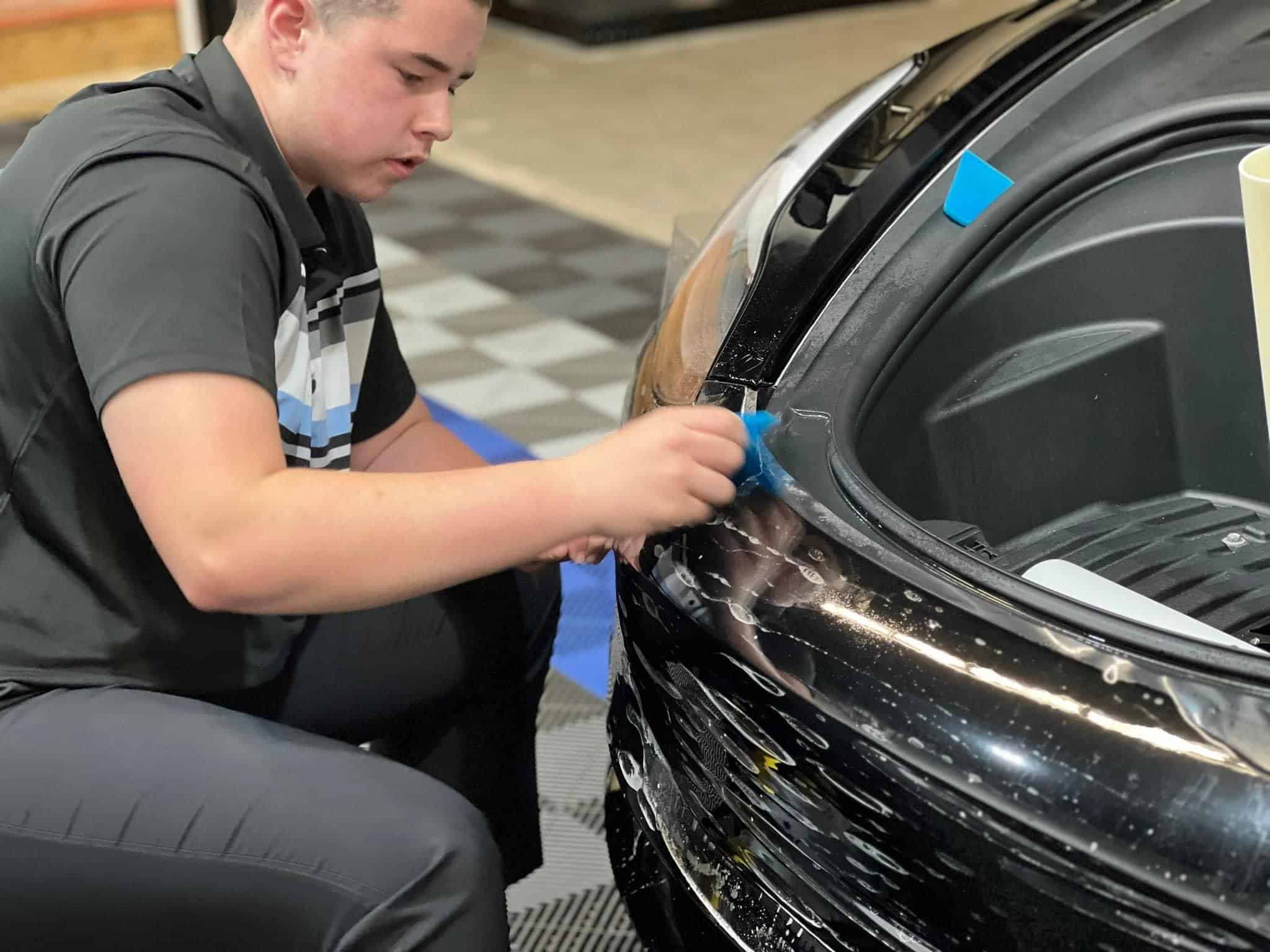 Technician smoothing blue protective film onto the front fender of a black car during detailing.