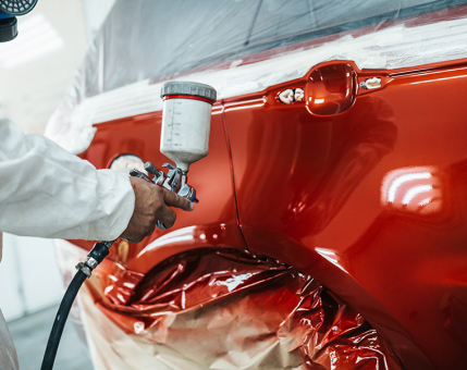 Person wearing protective gear paints a red car with a spray gun, masking tape and paper covering the area around the panel.
