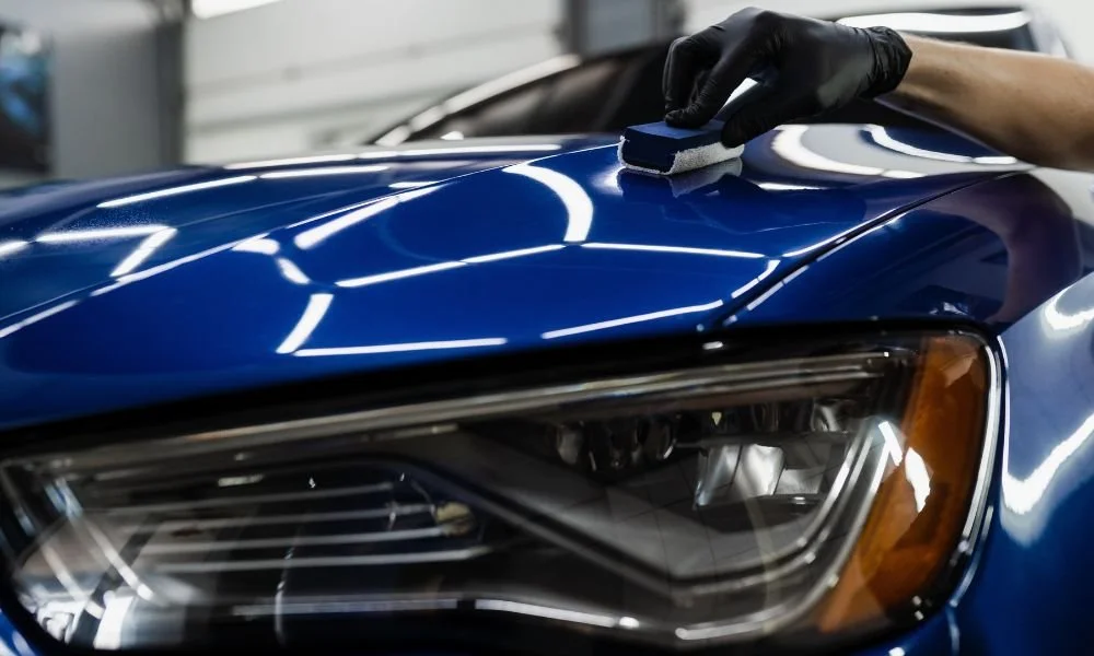 Gloved hand polishing a blue car hood with a sponge applicator.