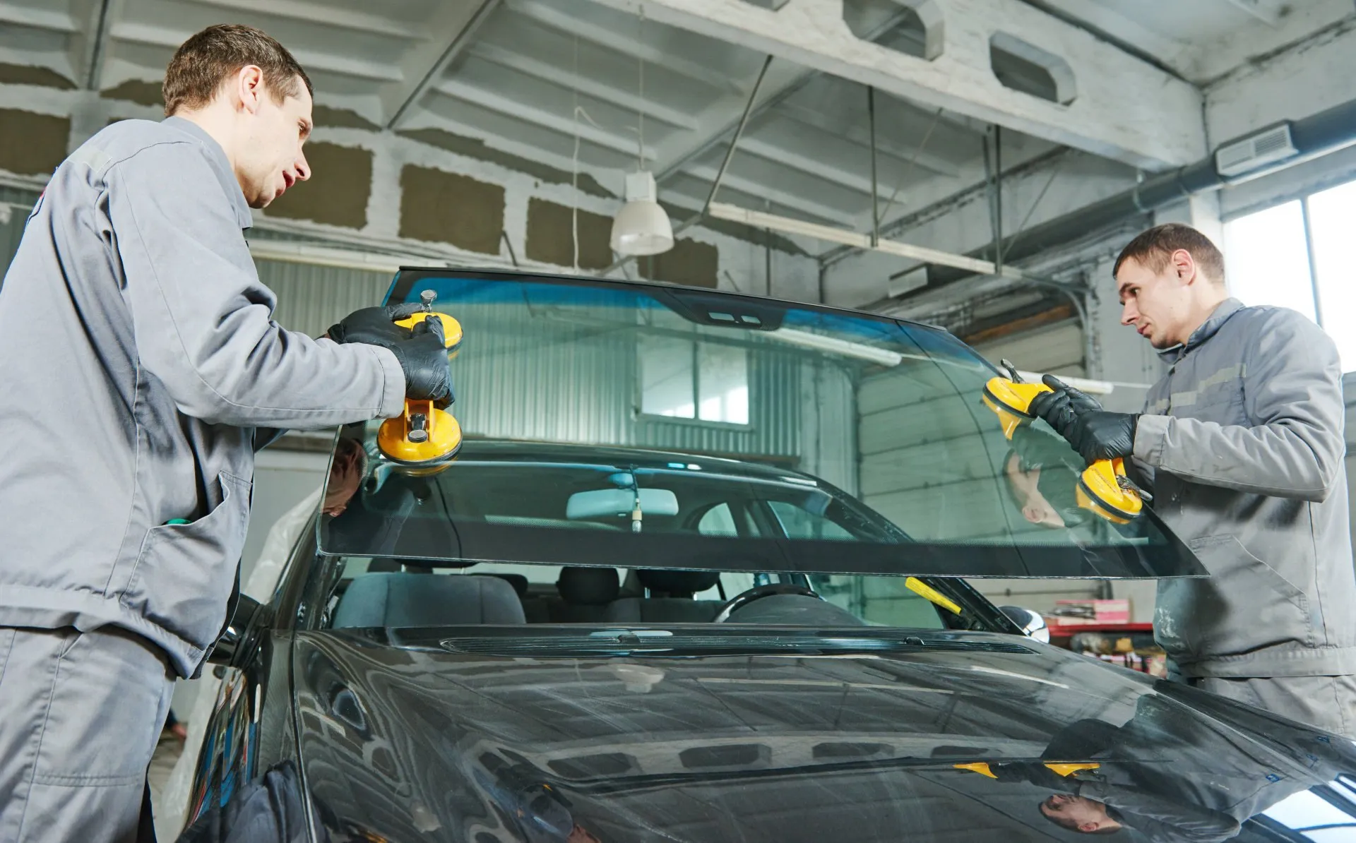 Two technicians in gray coveralls wearing gloves use yellow suction cups to install or remove a car windshield in a shop.