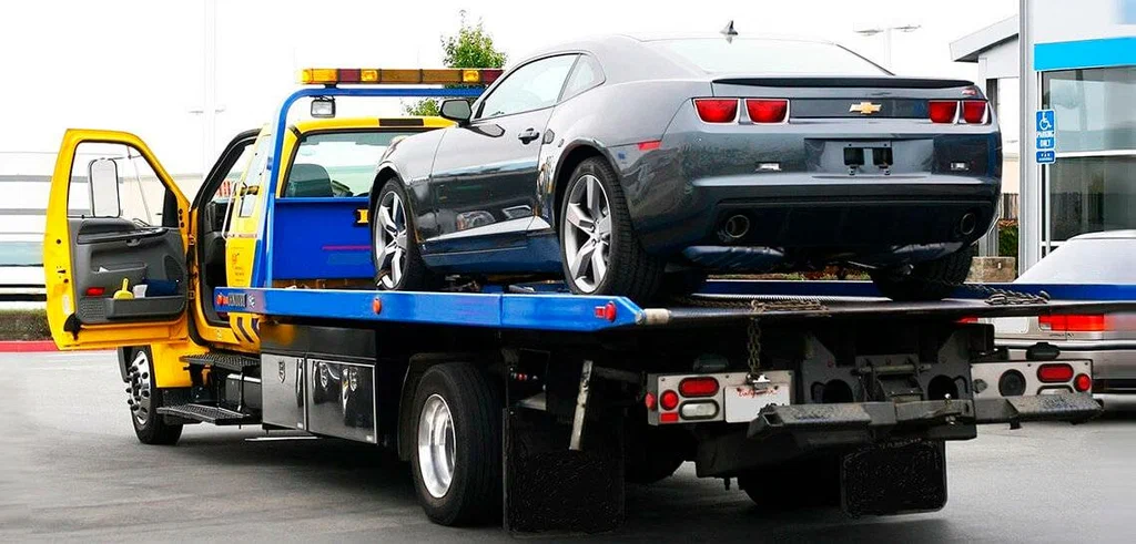 Yellow tow truck with its door open, transporting a gray Chevrolet Camaro on a flatbed trailer in a parking lot.