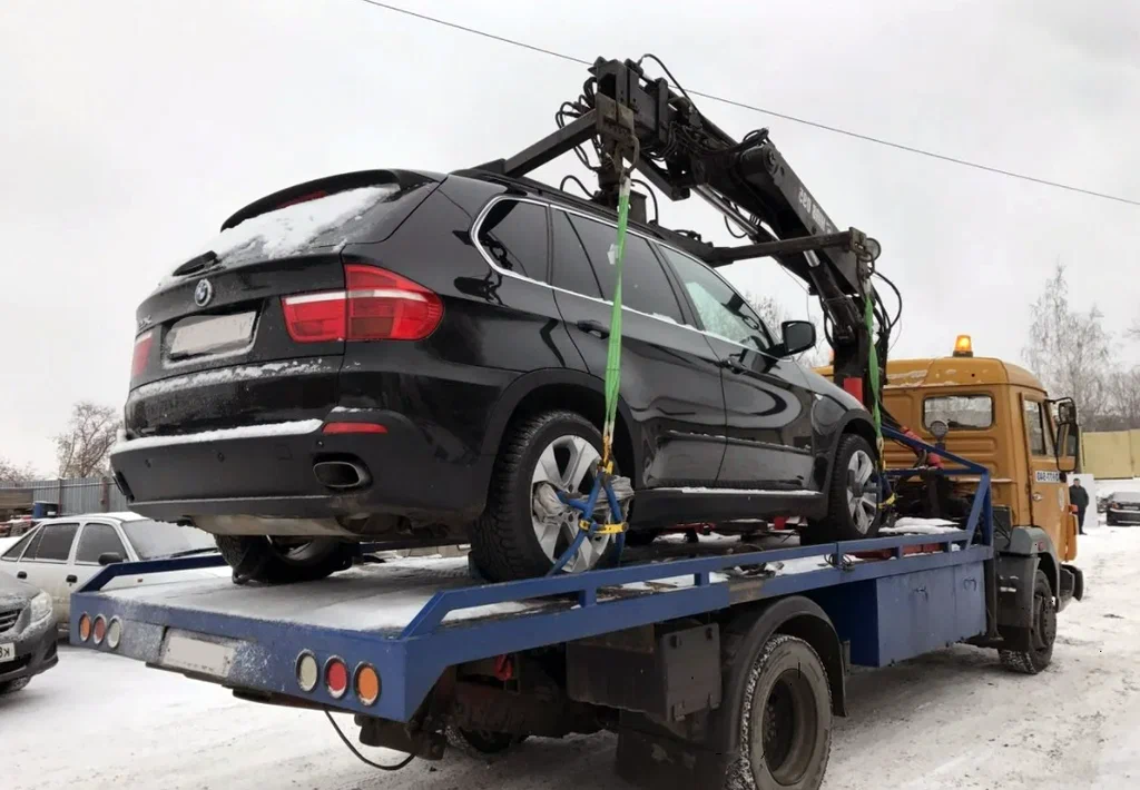 Black SUV secured on a blue flatbed tow truck with a crane overhead in a snowy lot.