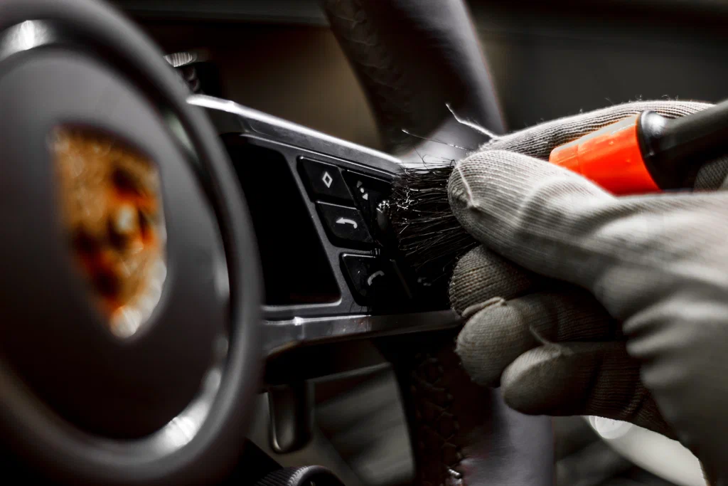 Close-up of a gloved hand cleaning car dashboard buttons with a brush.