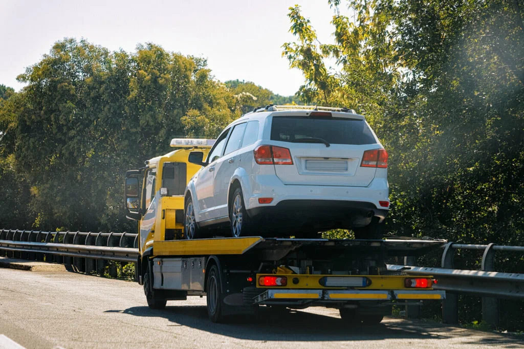 White SUV on a tow truck flatbed traveling on a sunny highway beside a guardrail.