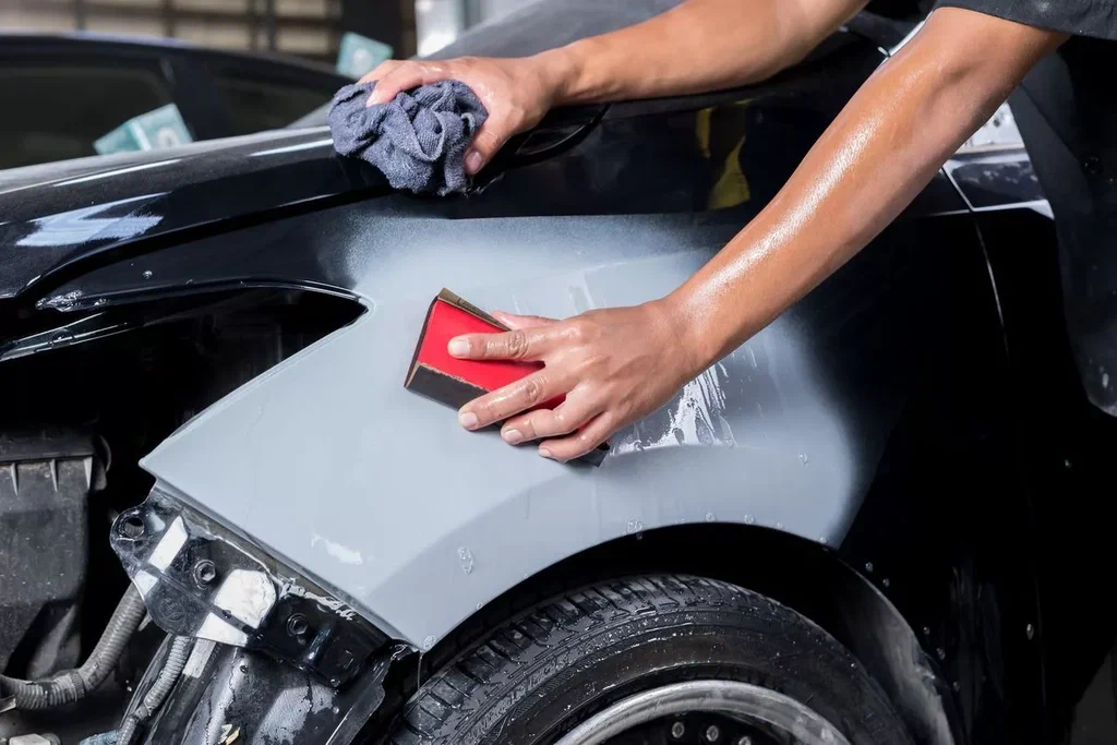Person detailing a car's front fender with a cloth and wax applicator, in a workshop setting