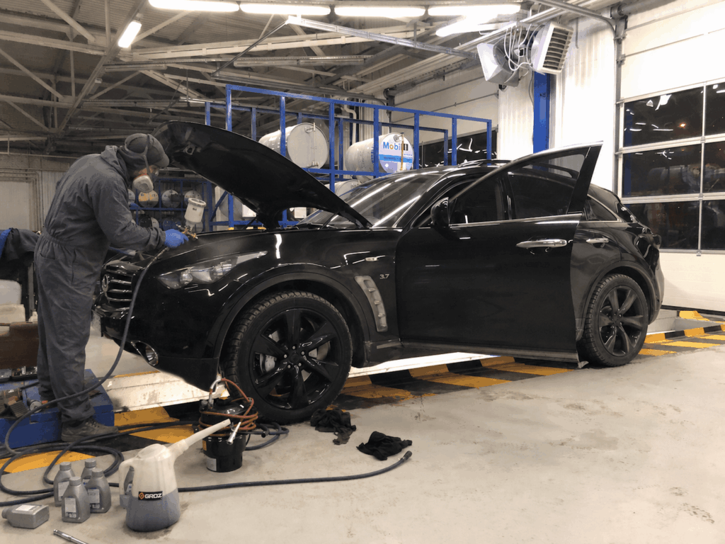 Technician in protective gear inspecting the engine with hood open on a black SUV in an auto shop of a repair facility.