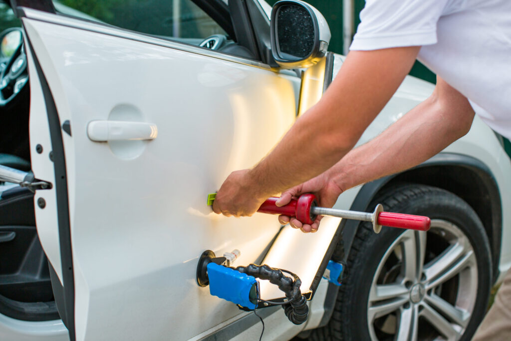 Person using a red unlocking tool to pry open a white car door manually, with jumper cables visible nearby and the door open.
