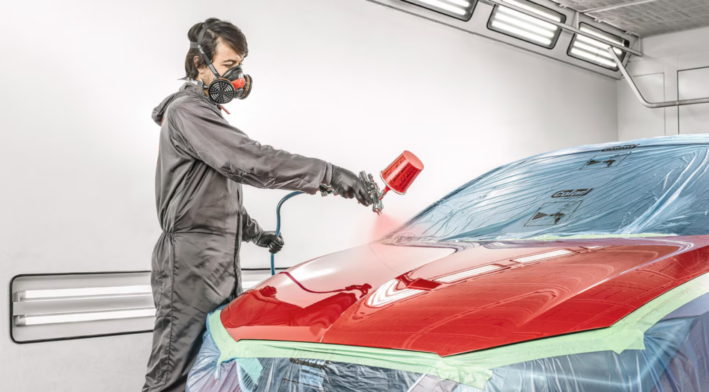 Auto body painter in a respirator mask spraying red paint onto a car in a spray booth.