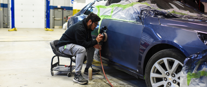 Mechanic polishing a blue car with a handheld buffer in a workshop, masking tape around the panels.