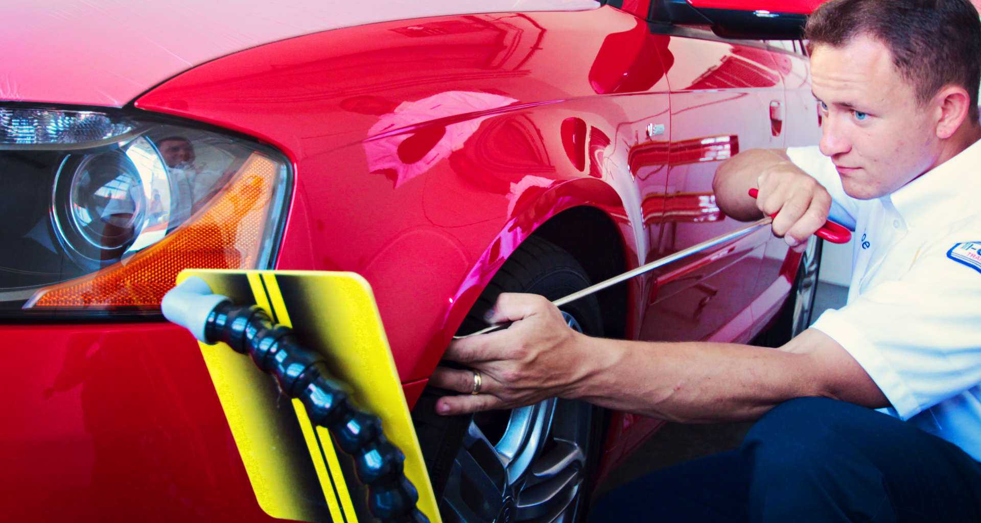 Automotive technician using a wheel alignment/diagnostic device on a red car's front wheel in a service bay.