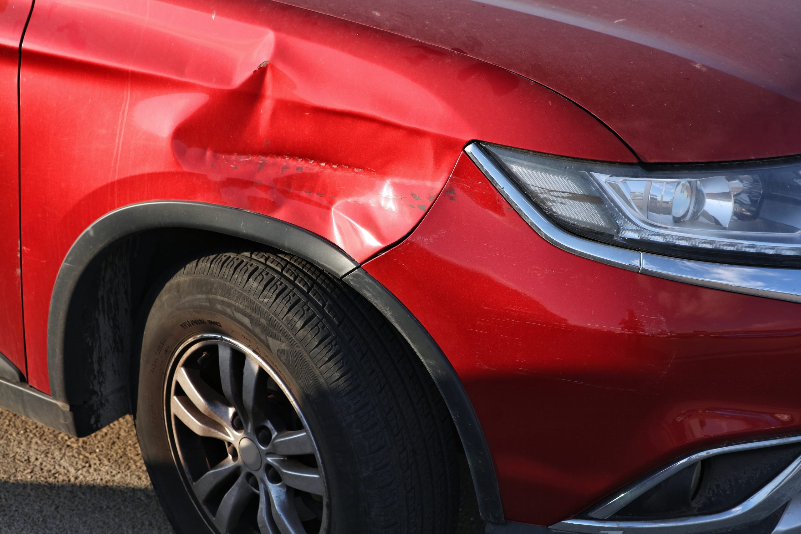 Red car with a large dent and scratches along the front fender near the headlight area, wheel visible below the damage
