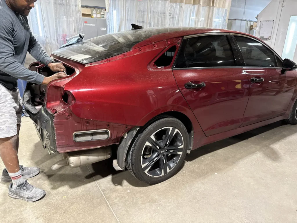 Technician repairs the rear area of a red car in a shop, with panels removed and tools in hand.