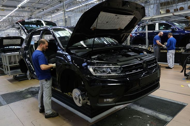 Automotive assembly line: a black SUV on a lift with its hood open and a technician inspecting the front wheel area.