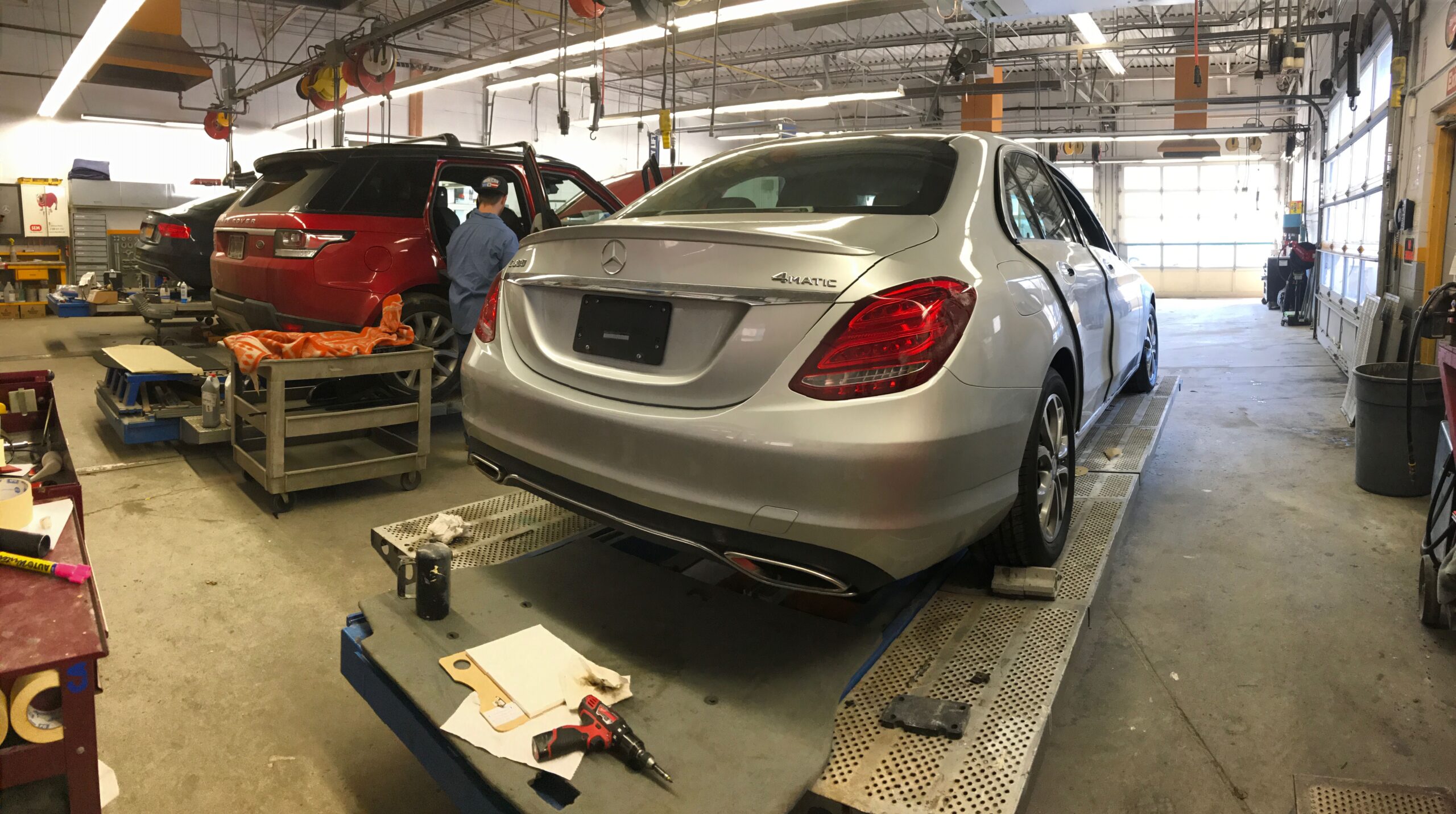 Auto repair shop with a silver Mercedes on a lift and a red SUV being serviced in the background; a technician works on the red car nearby.