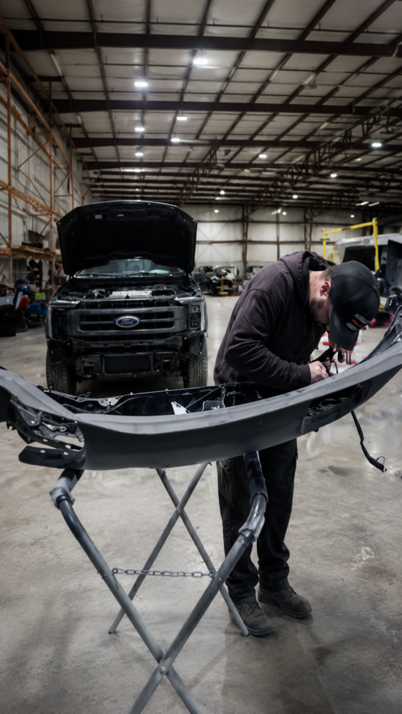 Auto technician in a cap and hoodie repairing a front bumper on a portable stand inside a large repair shop, with a Ford truck in the background under its hood.