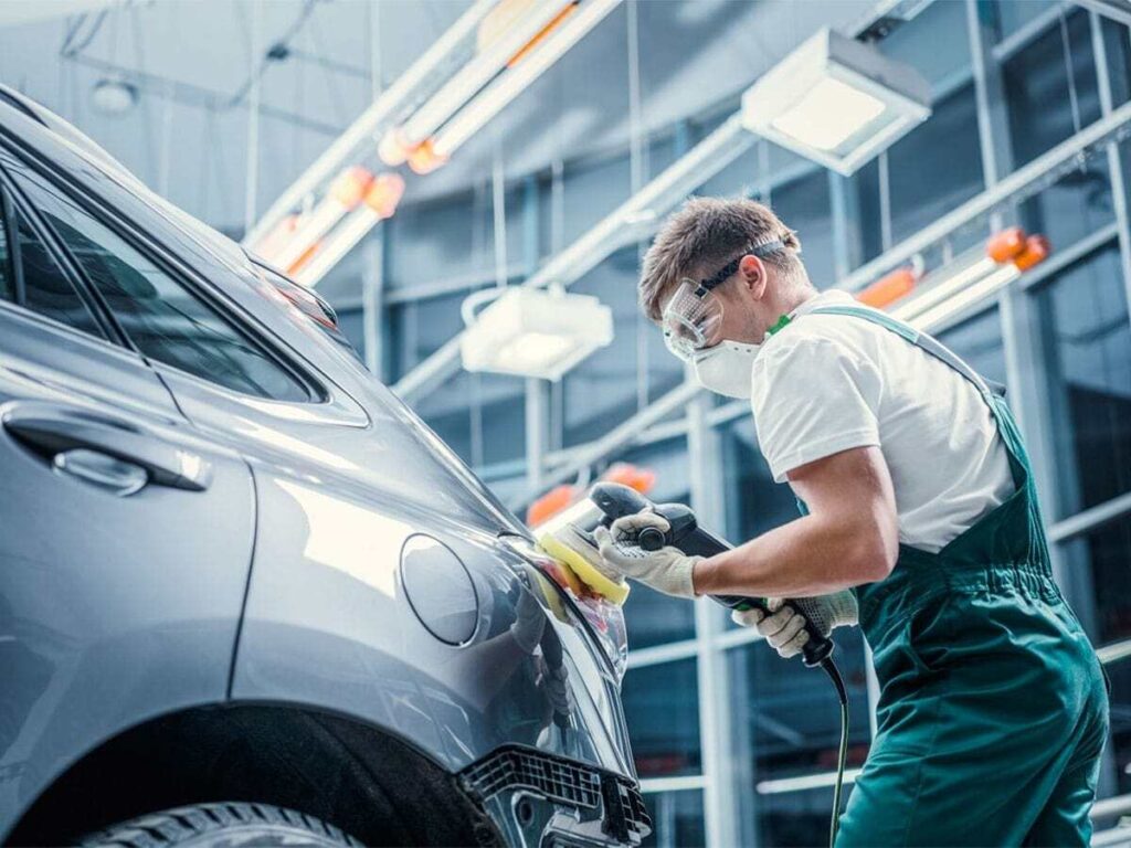 A maintenance worker wearing safety goggles and a face mask polishes the side of a silver car with a handheld buffer in a bright automotive workshop.